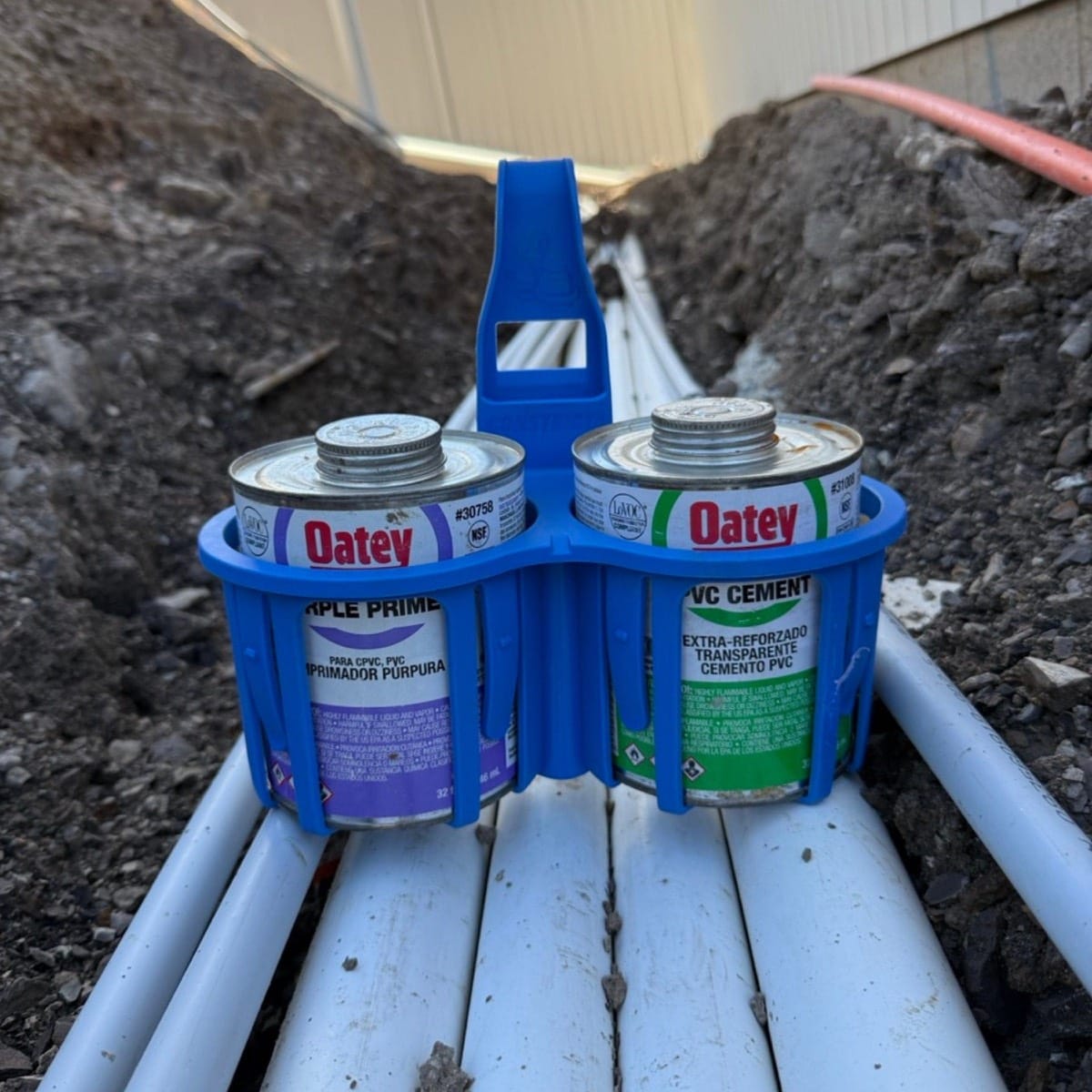 Two cans of Oatey paint and cement in a blue holder against a background of soil and concrete.